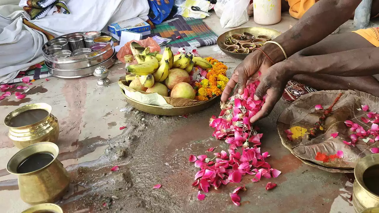 During a Hindu ceremony, hands scatter pink flower petals near offerings of fruit, marigolds, brass pots, and ritual powders.
