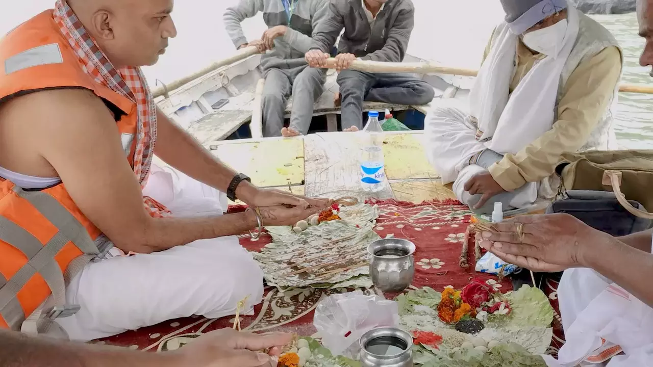 Men participate in a traditional ritual aboard a boat. Dressed in customary attire, with one wearing an orange life vest, they handle offerings like flowers and food placed on leaves. Another man rows the boat in the background during what appears to be a riverside ceremony.

