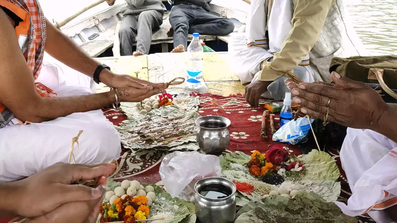 Men participate in a traditional ritual aboard a boat. Dressed in customary attire, with one wearing an orange life vest, they handle offerings like flowers and food placed on leaves. Another man rows the boat in the background during what appears to be a riverside ceremony - Pitrupaksha is in autumn