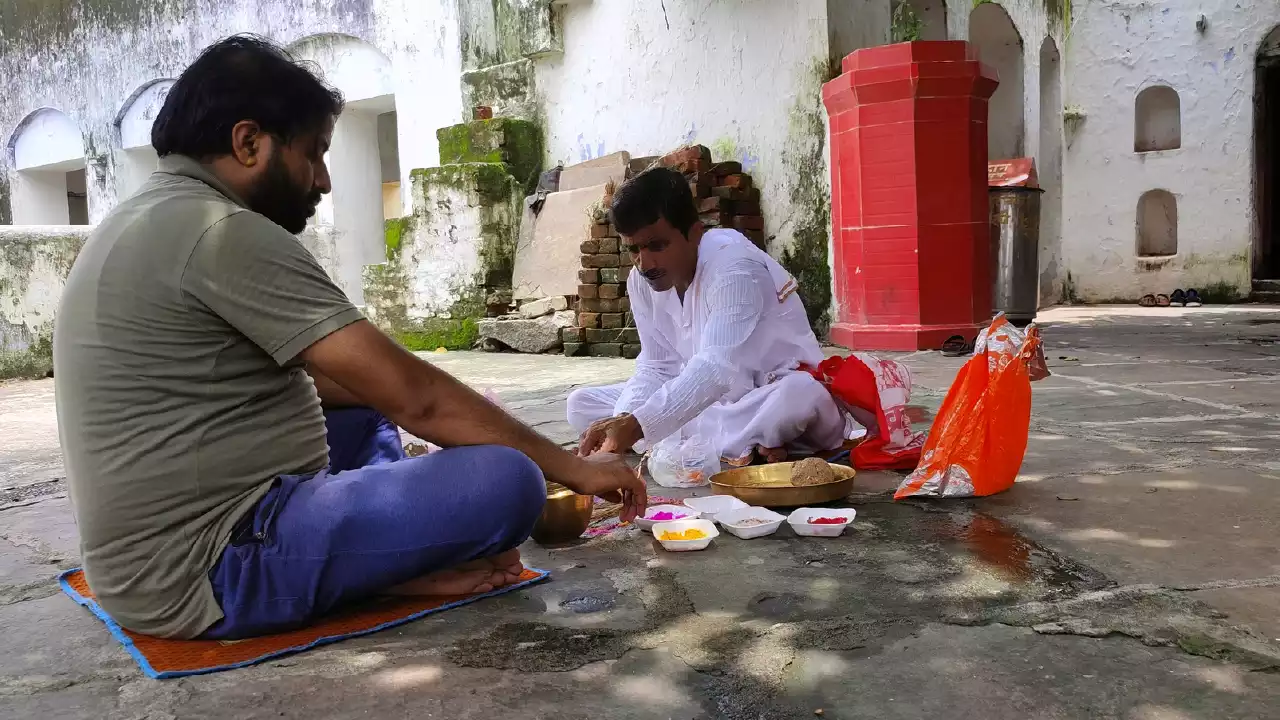 Image of shows two men sitting cross-legged on the ground, facing each other, outdoors in what looks like a courtyard or area near a building (possibly a temple). The man on the right is wearing white traditional clothing and appears to be leading or officiating a ritual. The man on the left wears a casual green t-shirt and blue jeans and sits on a small mat, participating. Between them are several small white bowls containing colorful powders (like yellow, pink, red) and other items, possibly including a brass bowl, used for the ceremony. The background features weathered white walls, some exposed brickwork, and a distinct red-painted structure to the right - Maha Bharani Pitrupaksha