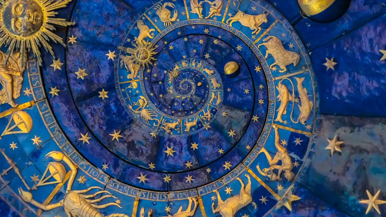 The image shows a detailed illustration of a zodiac wheel or astrological chart designed as a spiral. It's rendered in rich, deep blues and accented with elaborate gold details. Golden representations of the twelve zodiac signs (like Aries the ram, Taurus the bull, Gemini the twins, etc.) are clearly visible, arranged along the spiral path. Numerous golden stars are scattered across the blue background. A prominent golden sun symbol is visible towards the outer edge, and another smaller golden sphere (perhaps the moon or a planet) is near the center. The overall impression is one of ancient astronomy, astrology, or a celestial clock face - Maha Bharani Pitrupaksha