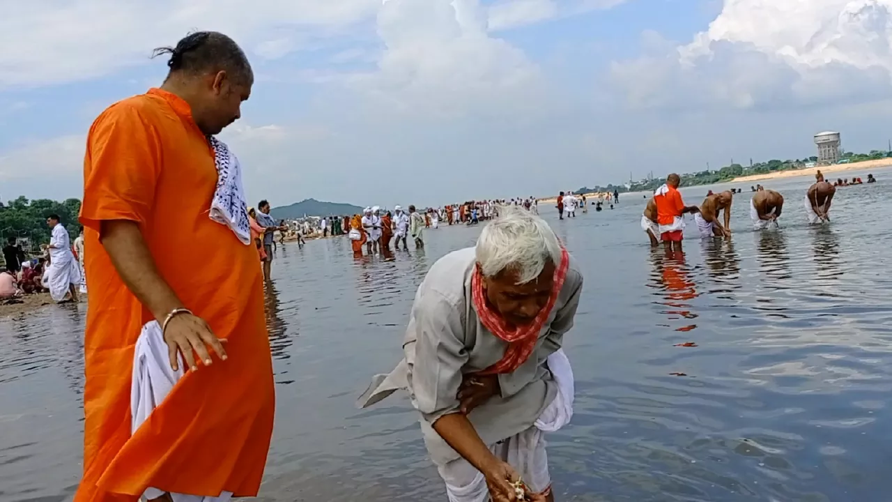 Elderly man performs Tarpan in river, offering water & grass from hands. Another man stands nearby. 