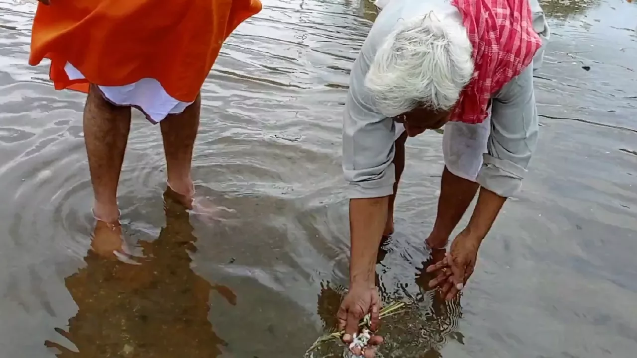 Image of Elderly man performs Tarpan in river, offering water & grass from hands. Another man stands nearby- Water symbolism Tarpan
