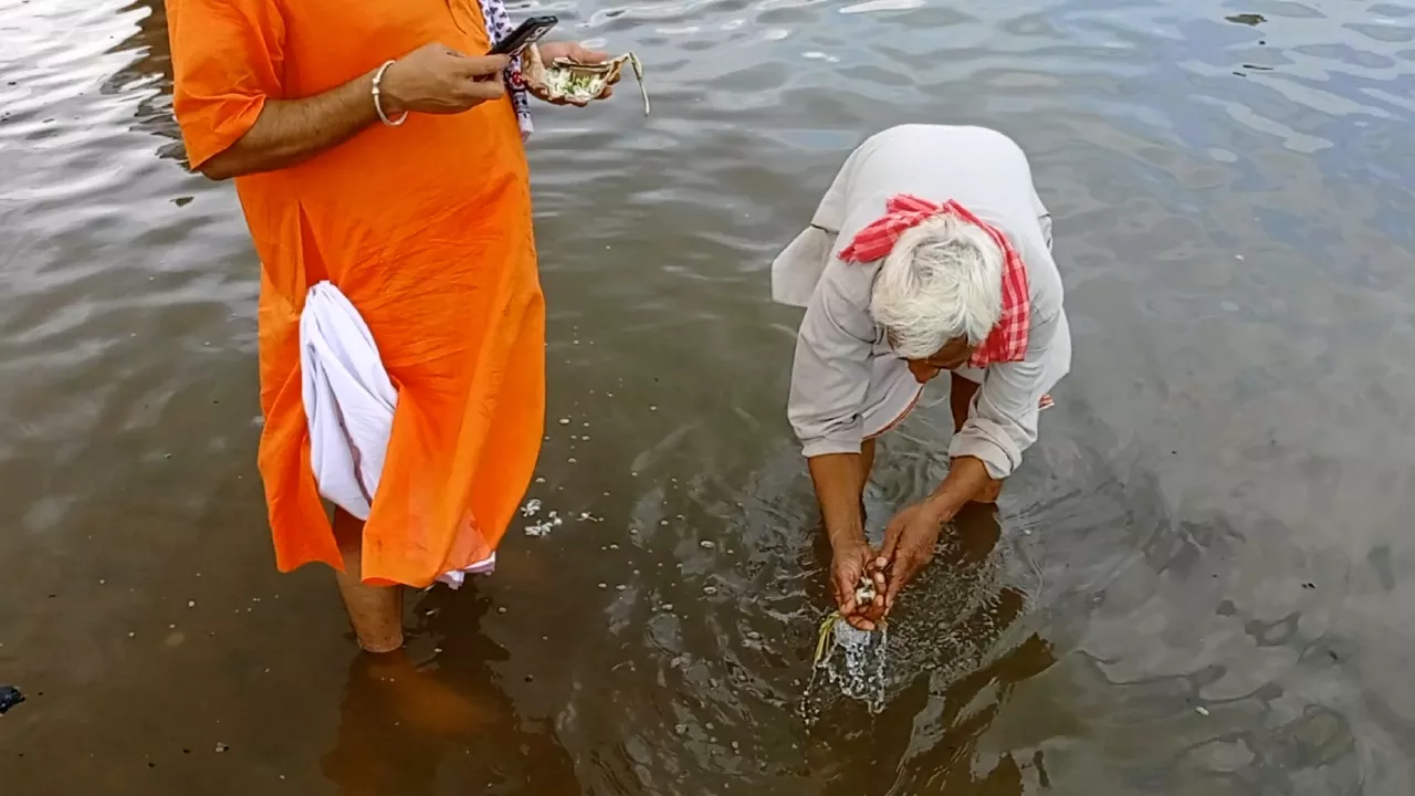 Image of Elderly man performs Tarpan in river, offering water & grass from hands. Another man stands nearby- Water symbolism Tarpan