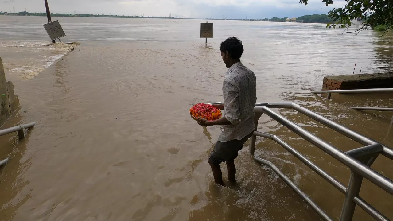 Man carries a plate of flower petals into a flooded river, standing on submerged ghat stairs.

