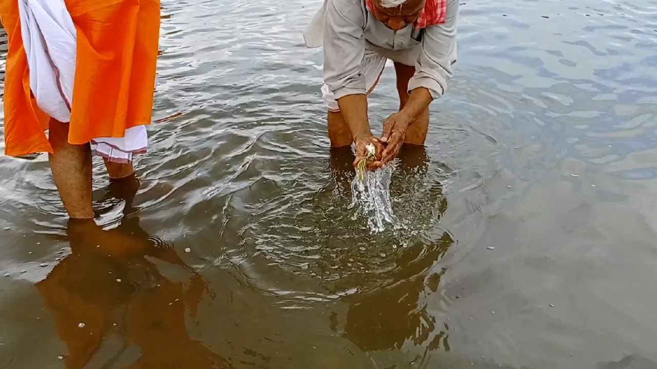 Image of Elderly man performs Tarpan in river, offering water & grass from hands. Another man stands nearby- Tarpan for Unknown Ancestors