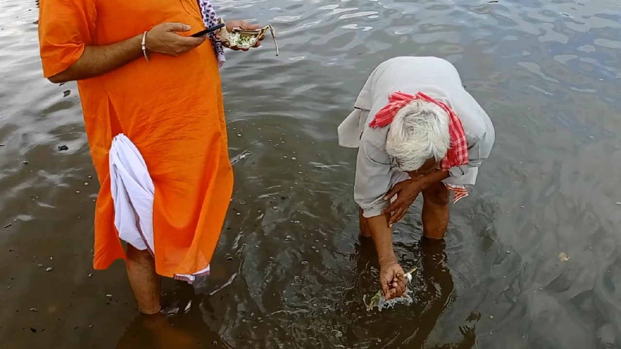 Images of Two men in river. Elderly man performs Tarpan ritual, offering water. Other holds items- Tarpan for maternal ancestors